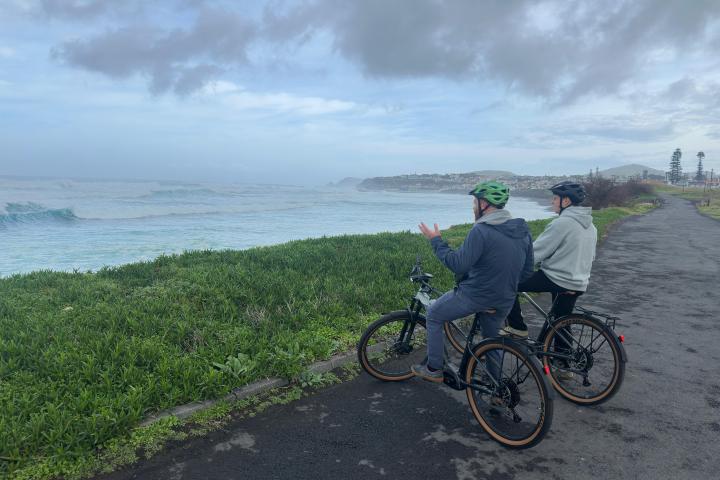 Two cyclists in helmets stop to view ocean waves from a coastal path on a cloudy day.