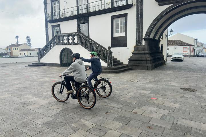 Two people riding bikes in a plaza near a historic building with an arched entrance.