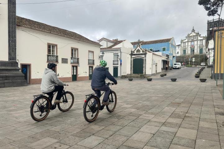 Two cyclists ride through a paved plaza in a small town with historic buildings.