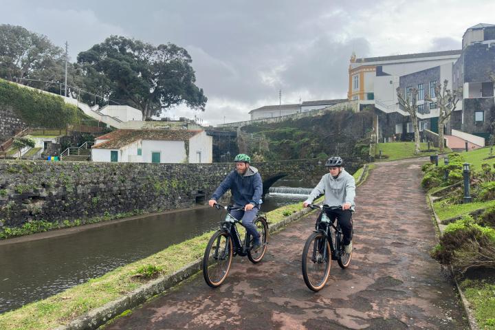 Two people biking on a path beside a canal with buildings in the background.