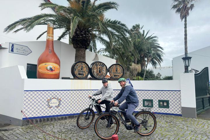 Two people on bikes in front of a large bottle and barrels, with palm trees and tiled wall in the background.