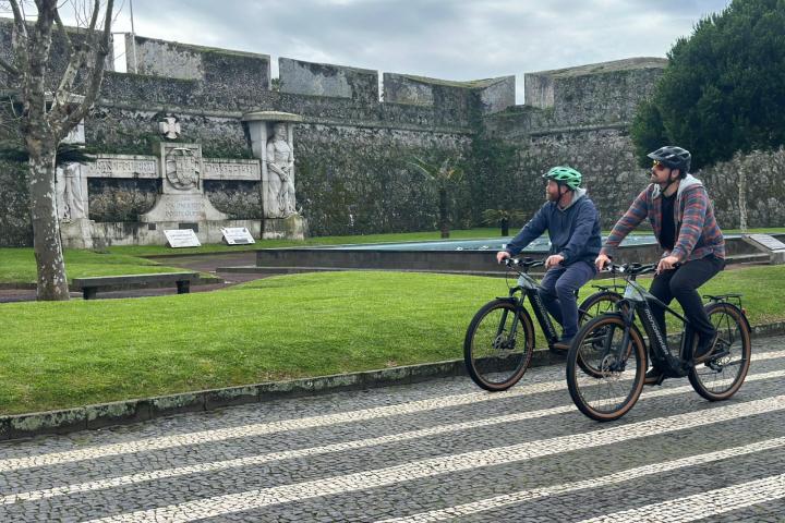 Two people on bicycles riding past a historical stone wall in a park.