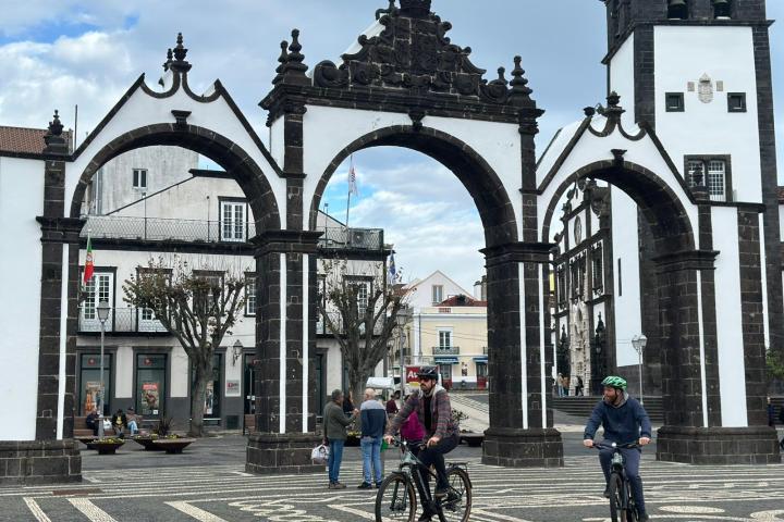 Two cyclists ride through historic stone arches in a city square, with people and buildings in the background.