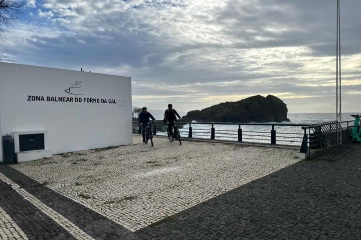 Two cyclists ride near a coastal area with rocky island and sign 'Zona Balnear do Forno da Cal'.