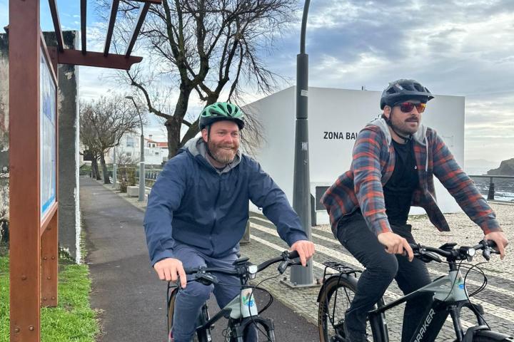 Two people biking on a path near trees and a building under a cloudy sky.
