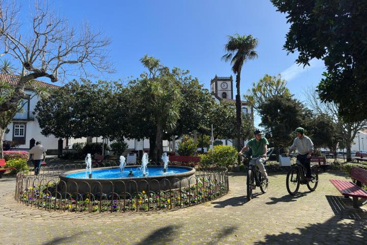 Two cyclists ride past a fountain in a sunny park with a clock tower in the background.