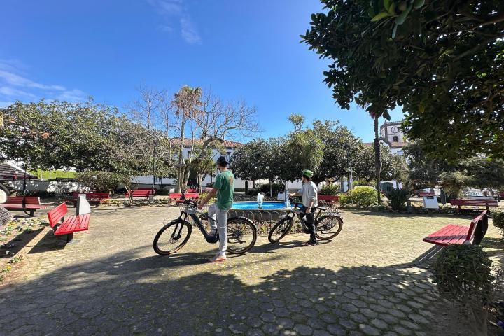Two people with bikes in a sunny park with red benches and trees.