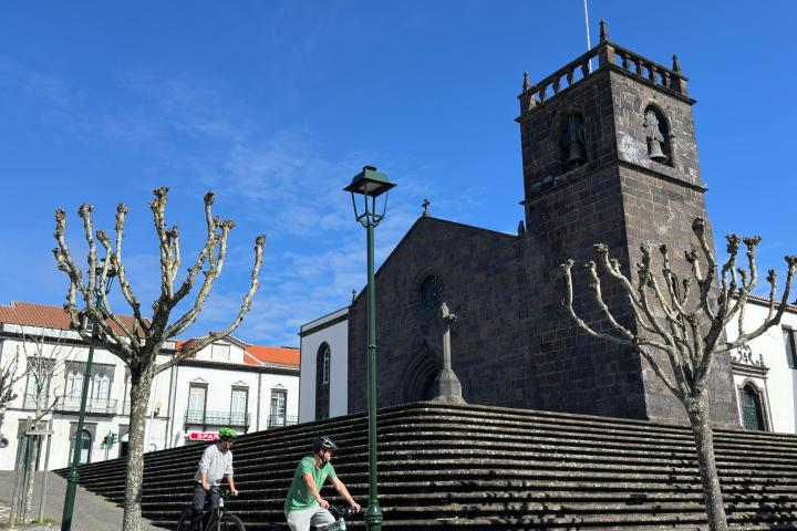 Two cyclists ride by a stone church with a bell tower on a sunny day.