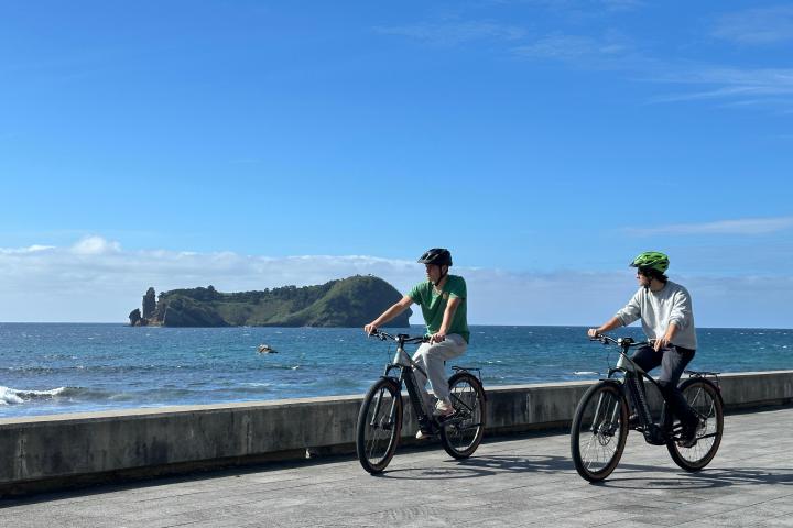 Two people cycling by the sea with an island in the background under a clear blue sky.