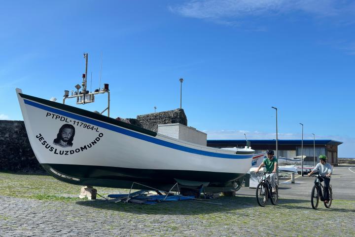 Two people on bicycles near a parked boat named Jesus Luz do Mundo on a sunny day.