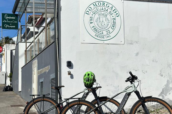 Two bicycles with a green helmet parked by a building with 'Do Morgado' sign on a sunny day.