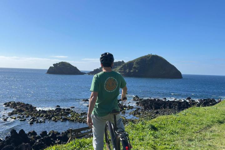 Person with bicycle stands on grassy cliff overlooking ocean and small islands.