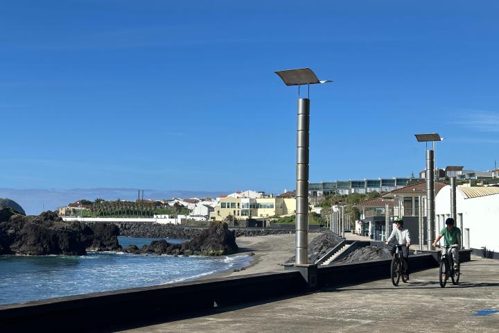 Two cyclists ride along a coastal path with ocean waves and buildings in the background.