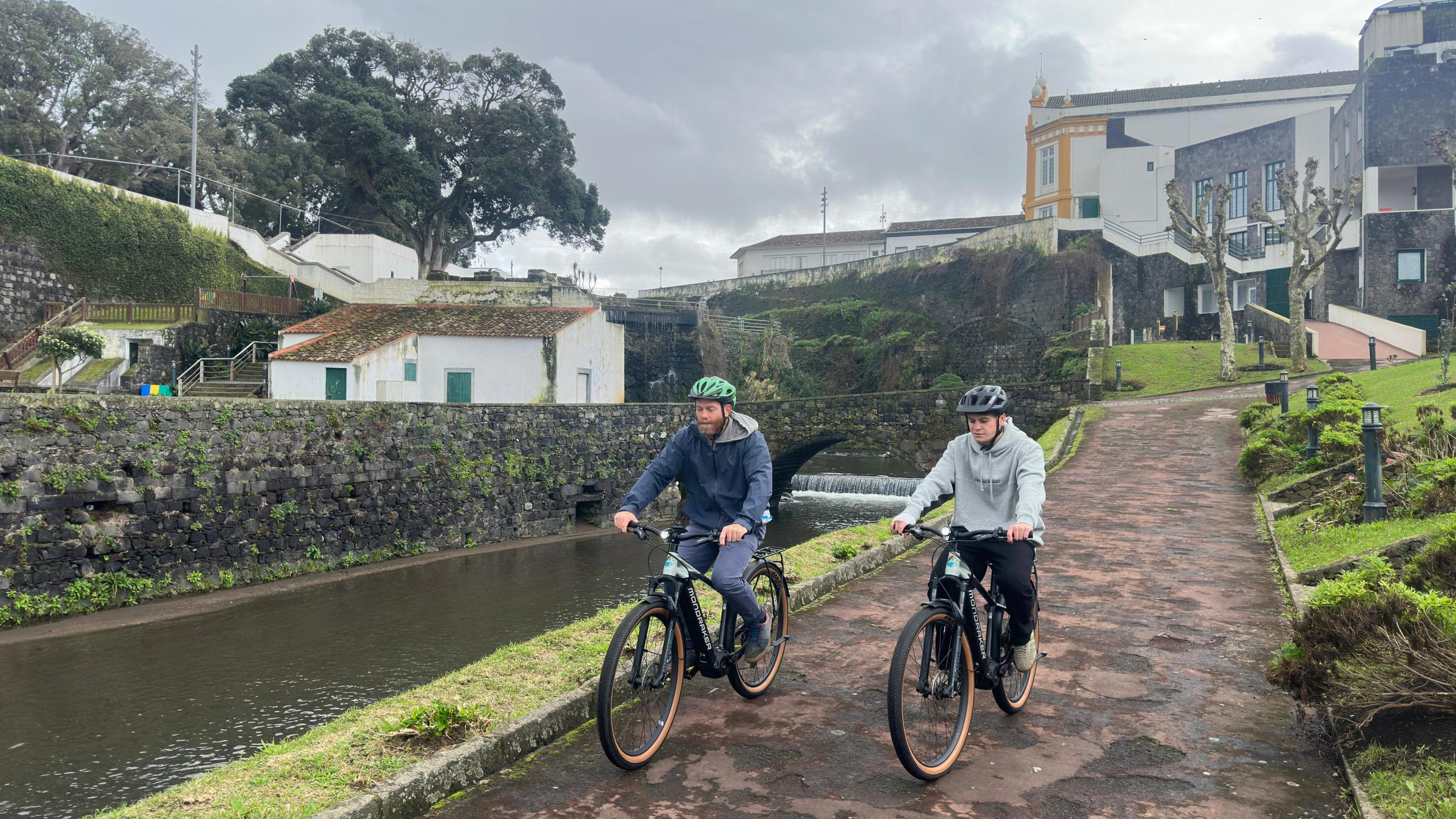 Two people cycling on a path by a river, with buildings and greenery in the background.