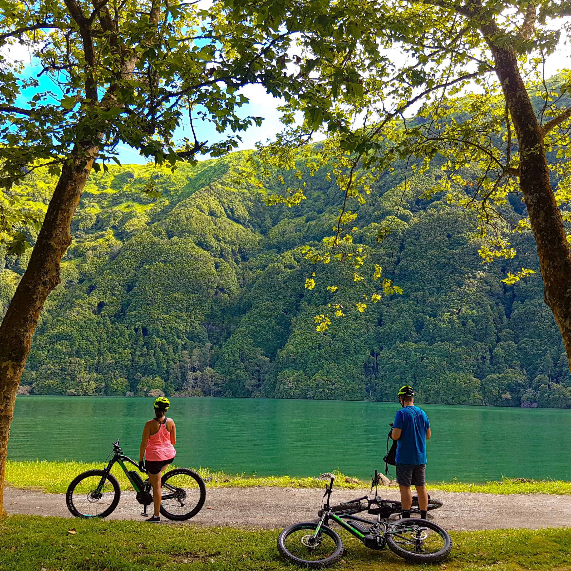 Two cyclists rest by a lake with lush green hills in the background.